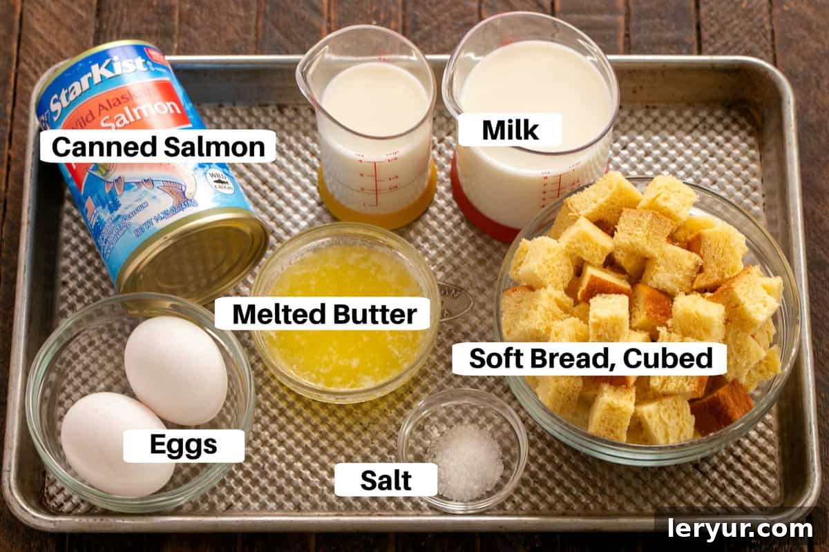 A baking sheet displays the simple, key ingredients for salmon loaf, neatly labeled for easy identification: eggs, milk, salt, a can of red salmon, and cubed brioche bread, ready for mixing.