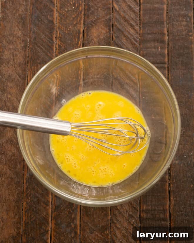 A close-up shot of two fresh eggs being vigorously whisked in a glass bowl, creating a light and airy foam.