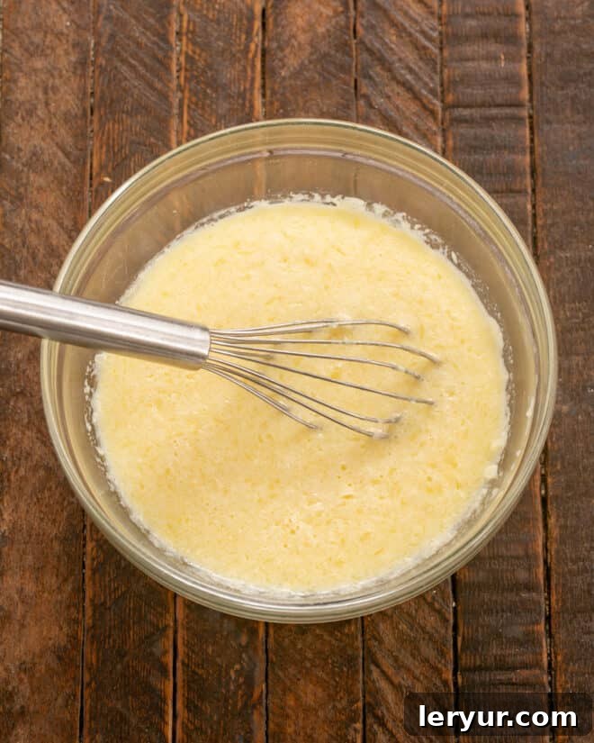 A hand stirring a creamy mixture of melted butter, milk, and salt into whisked eggs in a glass bowl.