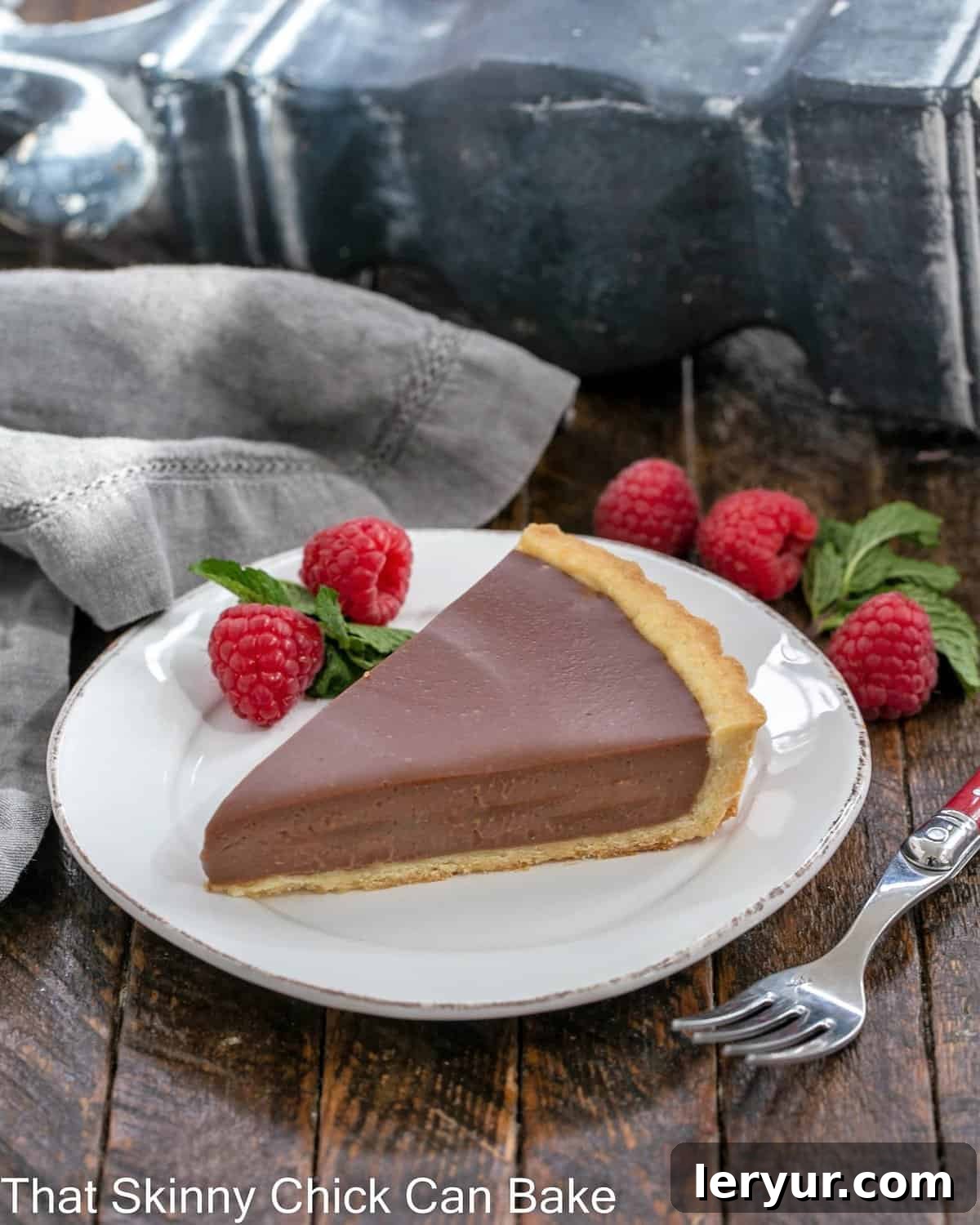 A perfectly sliced milk chocolate tart, garnished with a vibrant raspberry and a fresh mint leaf, rests on a crisp white plate.