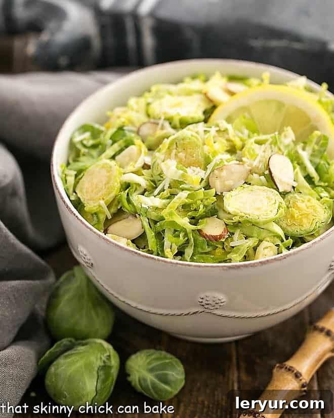 Crisp, refreshing Shaved Brussels Sprouts Salad with a lemony Dijon dressing - a healthy, delicious side dish! #healthy #salad #Brusselssprouts #lemon #almonds #Dijon #thatskinnychickcanbake Shaved Brussels Sprouts Salad in a white salad bowl with a lemon slice and a bamboo handled fork, on a rustic table.