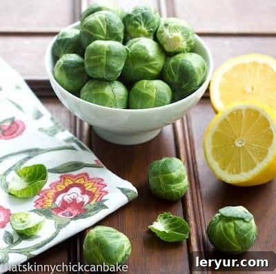 How about a raw Shaved Brussels Sprouts Salad with a lemony Dijon dressing? With Brussels sprouts being all the rage. This recipe is healthy and delicious! Brussels Sprouts in a small white bowl next to sliced lemons on a wooden table.