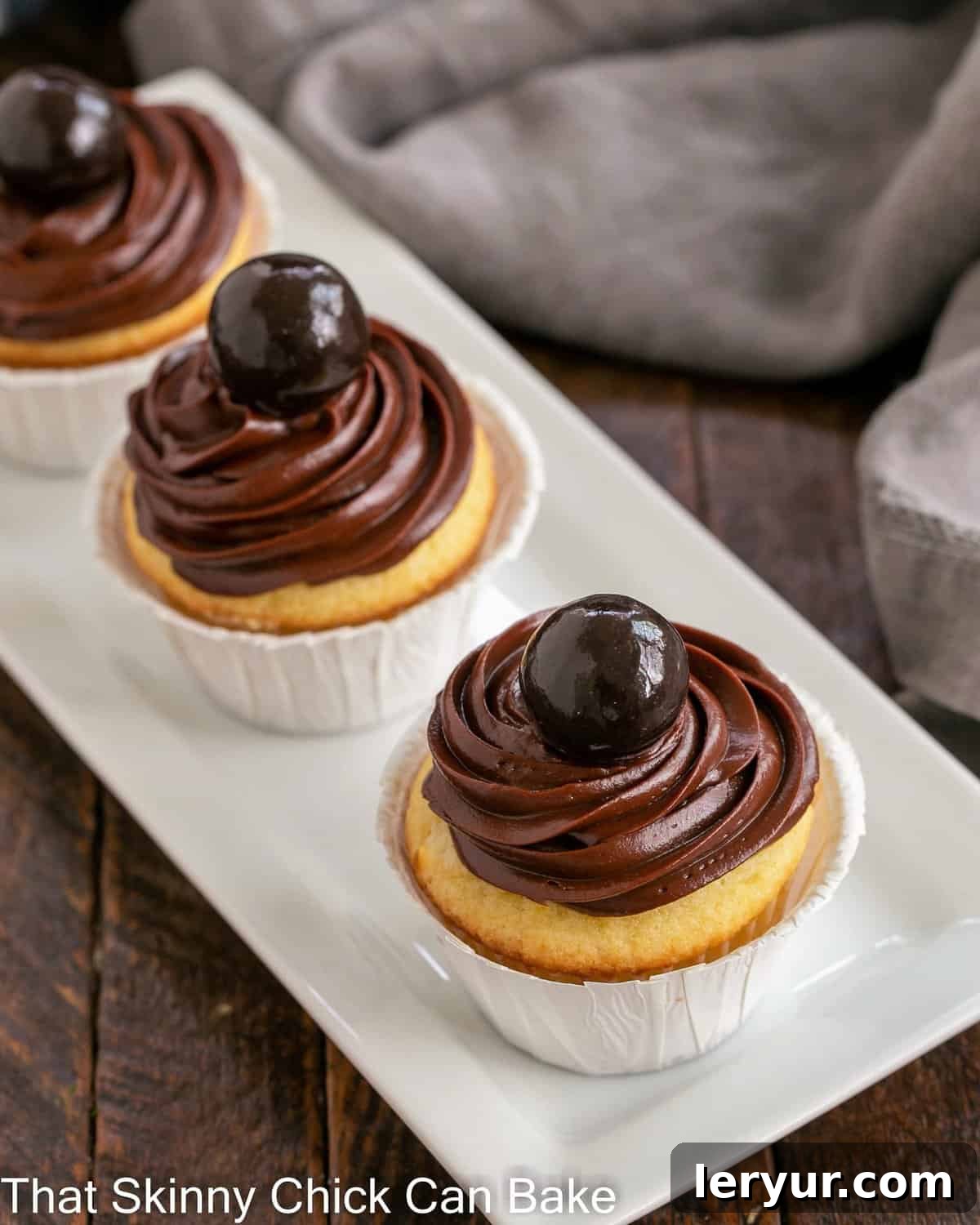 Golden Bliss: Cupcakes Crowned with Silky Ganache 2 Three beautifully frosted yellow cupcakes with chocolate ganache on a pristine white ceramic tray, ready to be enjoyed.