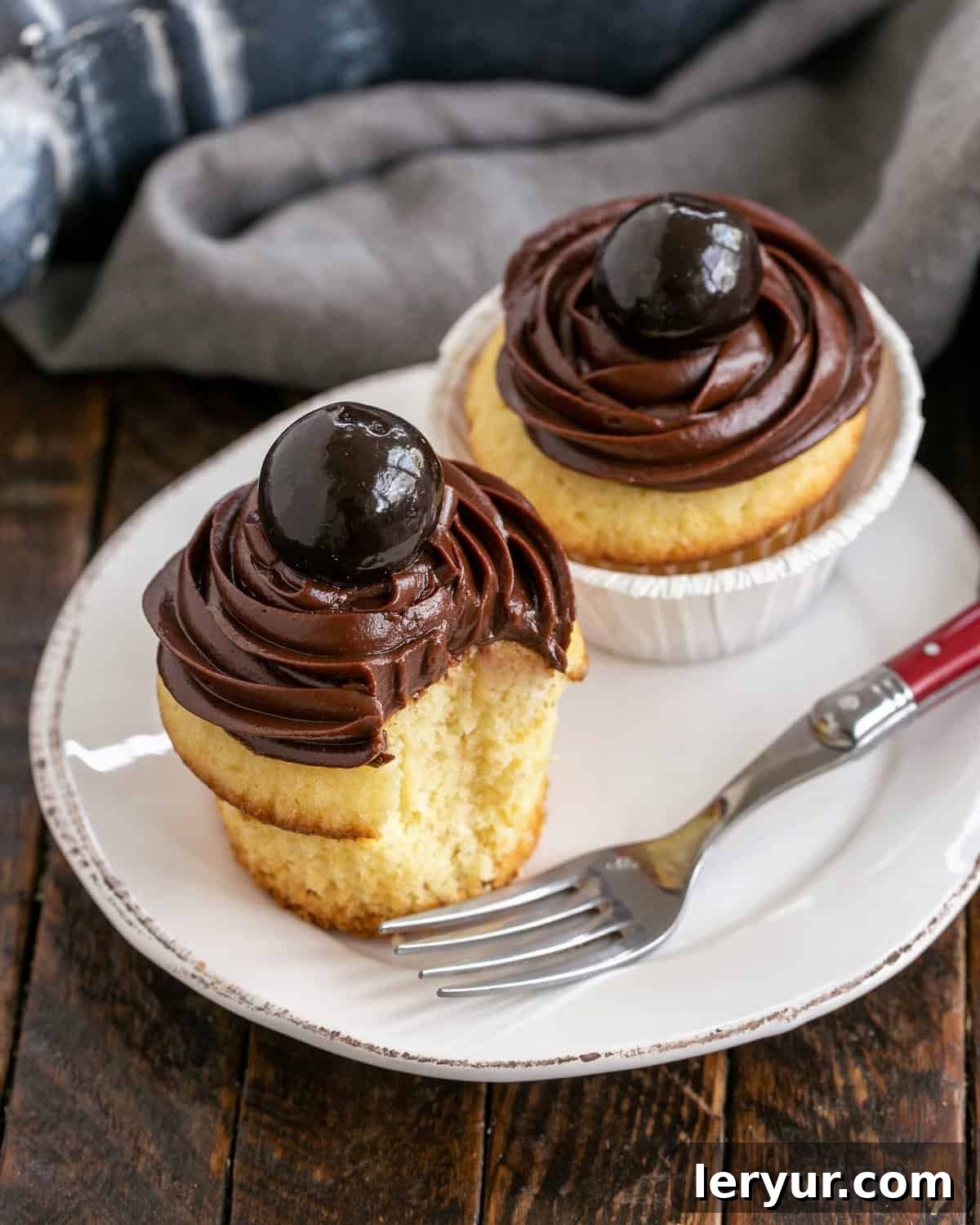 Golden Bliss: Cupcakes Crowned with Silky Ganache 3 Two golden yellow cupcakes elegantly frosted with chocolate ganache, presented on a white plate with a red-handled fork, inviting a taste.