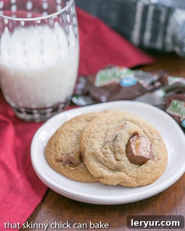 Two Milky Way Cookies with visible chunks of Milky Way candy on a small white plate, accompanied by a refreshing glass of milk.