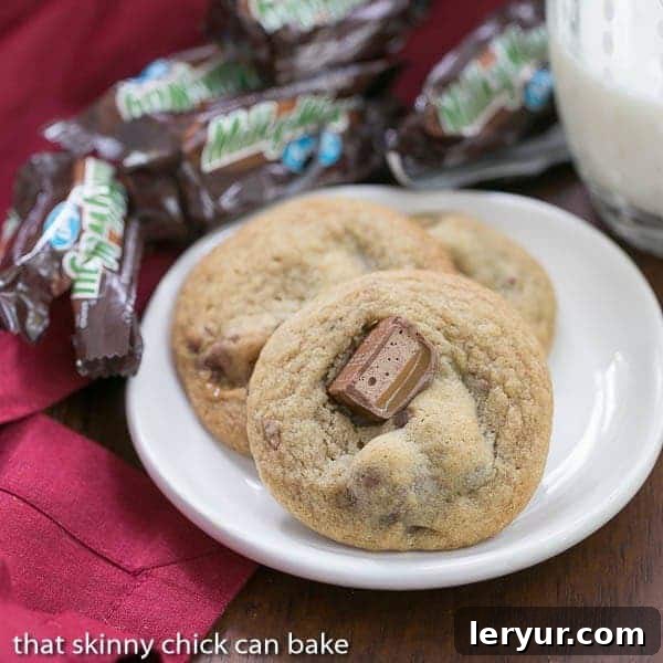 Three perfectly baked Milky Way Cookies arranged on a pristine white plate, surrounded by empty Milky Way candy wrappers, hinting at their delicious origins.
