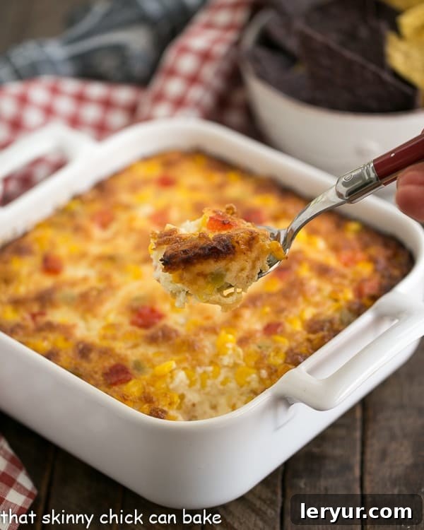 A serving spoon lifting a generous portion of warm, cheesy Fiesta Corn Dip from a white casserole dish, showcasing its gooey texture.
