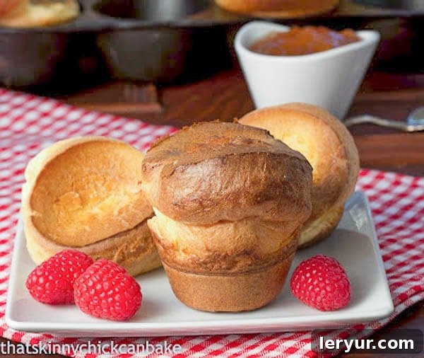 Perfect Popovers - puffed, eggy muffins that are a marvelous addition to your dinner menu, offering a delightful texture and taste. Freshly baked Perfect Popovers served on a square white plate, garnished with vibrant fresh raspberries for a touch of sweetness.