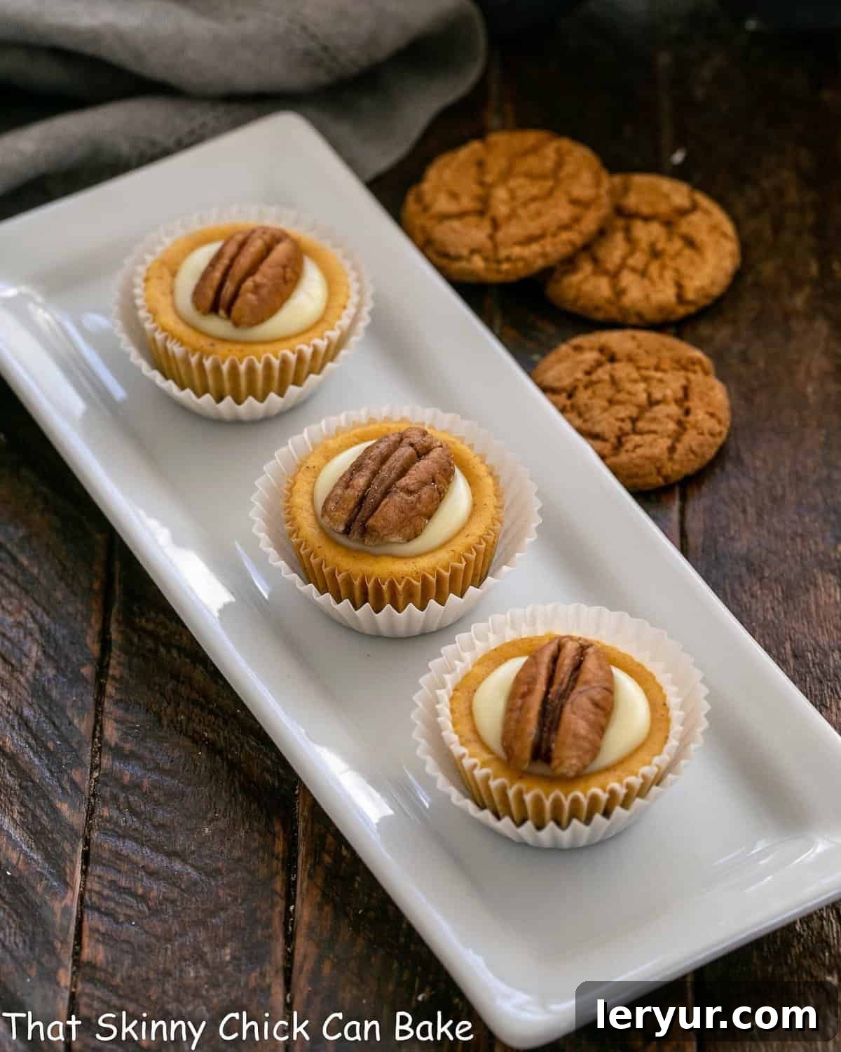 Three perfectly crafted mini pumpkin cheesecakes arranged artfully on a pristine white tray, with a backdrop of rustic ginger cookies suggesting the inviting crust.