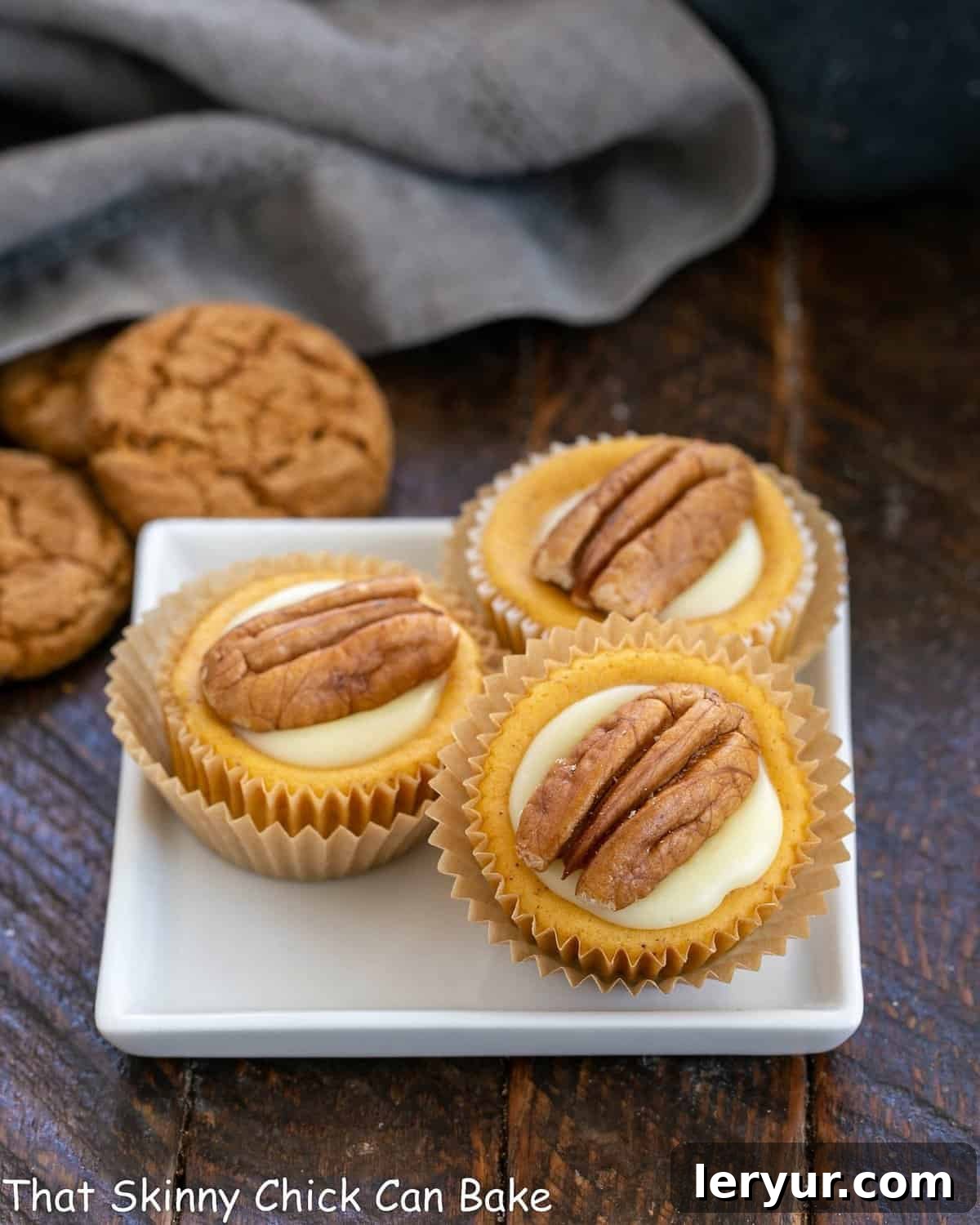 Three perfectly baked mini pumpkin cheesecakes, elegantly arranged on a square white plate, each topped with a delicate swirl of white chocolate ganache and a single pecan half.