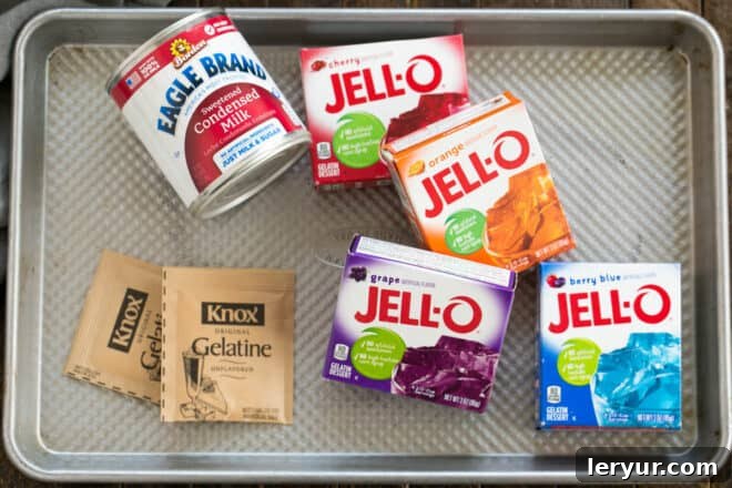 The essential ingredients for Broken Glass Jello, including flavored gelatin boxes, unflavored gelatin, and sweetened condensed milk, neatly arranged on a baking sheet.