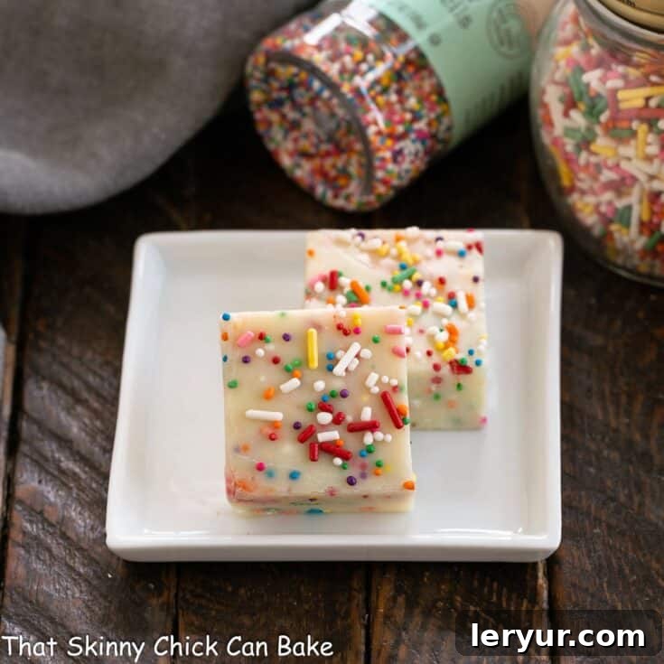 Two squares of white chocolate fudge on a white plate with jars of sprinkles in the background.