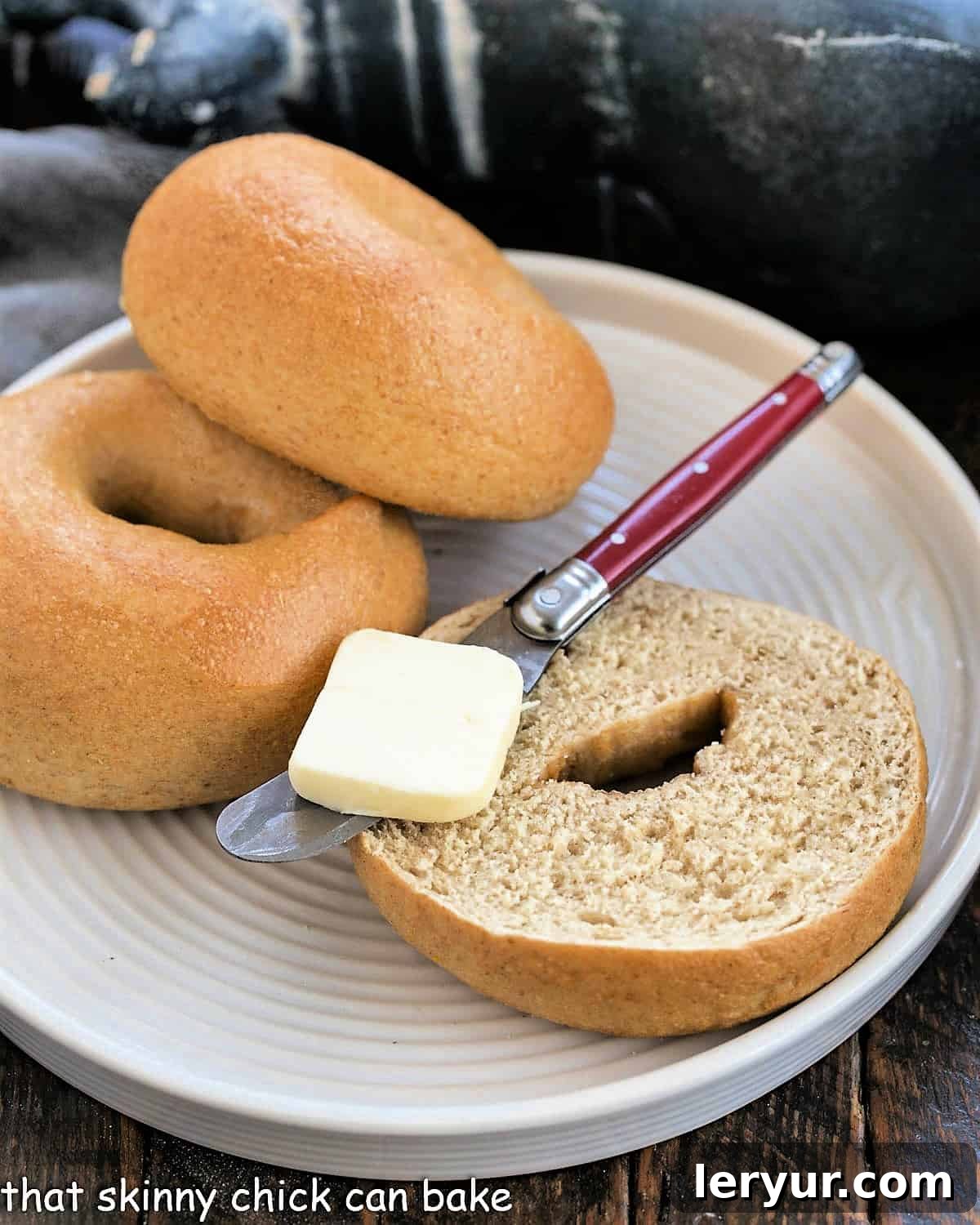 Two golden-brown whole wheat bagels on a white plate, one sliced open with a knife and a pat of butter, showcasing its dense, chewy interior.