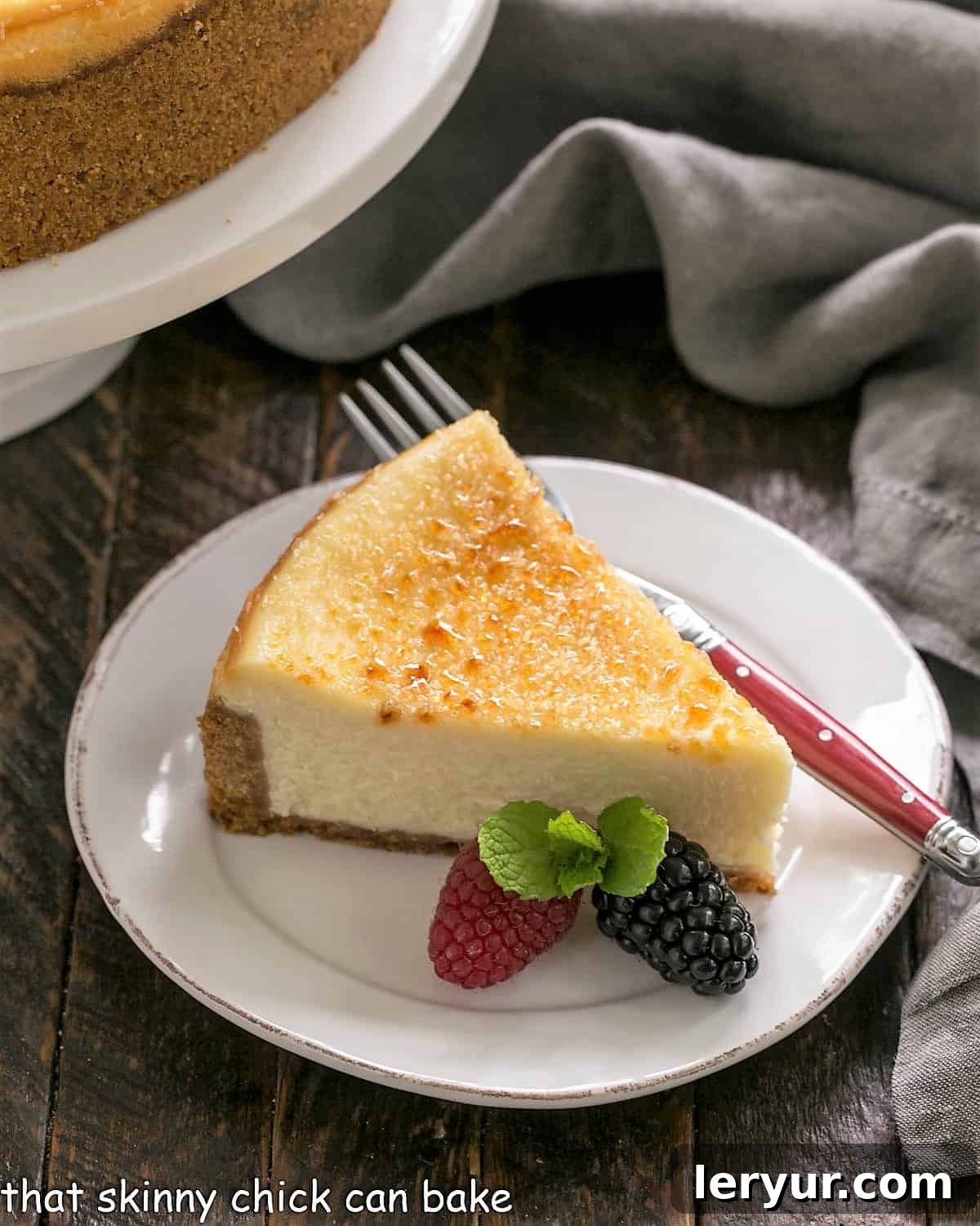 Crème Brûlée Cheesecake - a cross between 2 delicious desserts for a magnificent cheesecake recipe Slice of creme brulee cheesecake on a white dessert plate with a red handle fork, two berries and a sprig of mint.