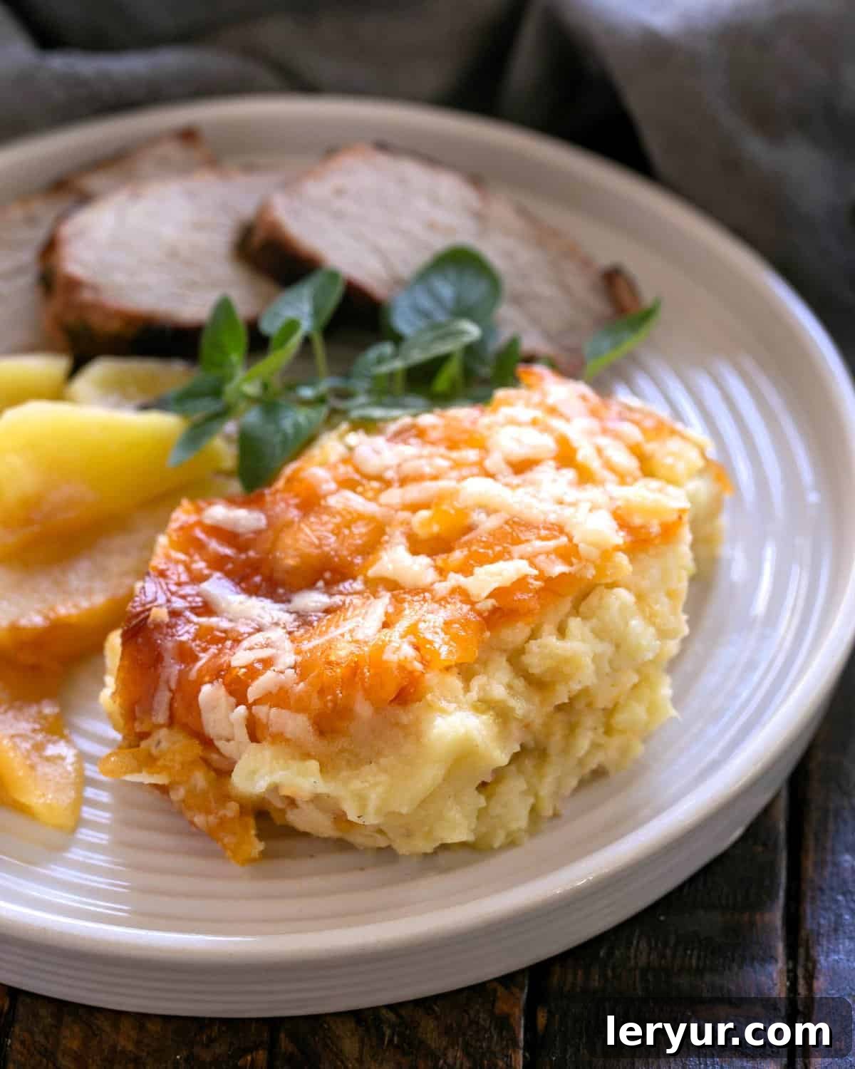 A delicious serving of twice baked potato casserole on a white dinner plate, garnished with fresh green onions, alongside roasted pork and apples.