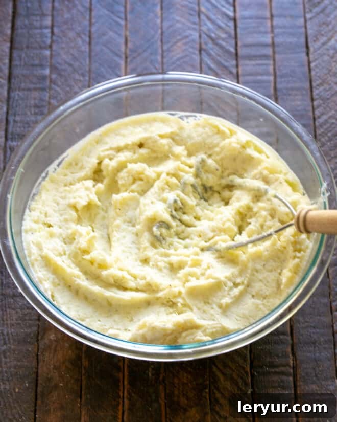 Mashed boiled potatoes being mixed with sour cream, heavy cream, salt, and pepper in a large mixing bowl.