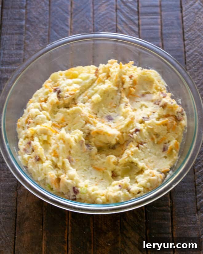 The potato mixture, now combined with bacon, cheese, and green onions, being spread into a casserole dish after an egg has been incorporated, ready for a final cheese topping.