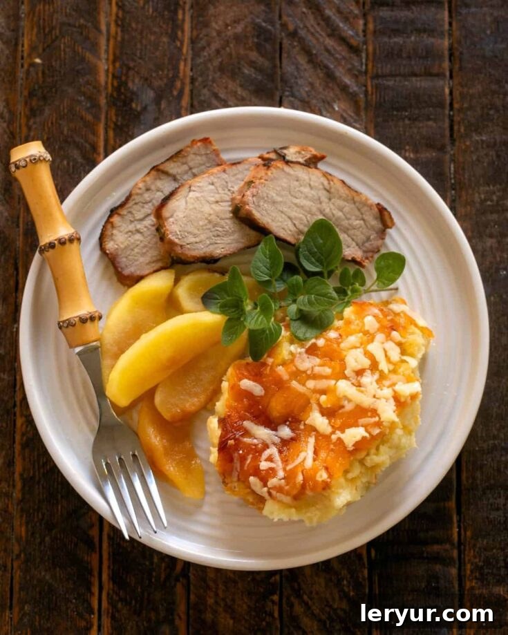 Overhead view of a golden-brown Twice Baked Potato Casserole, served on a white plate with roasted pork and apples, ready to be enjoyed.