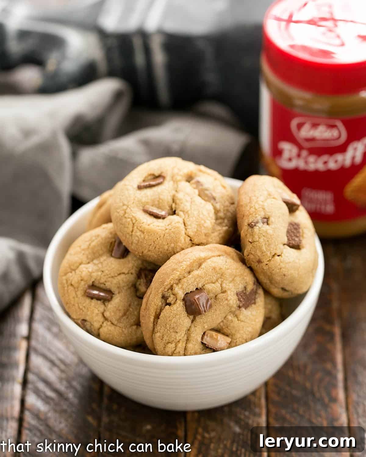 A white bowl filled with golden-brown Toffee Biscoff Cookies, with a jar of Biscoff cookie butter subtly blurred in the background, suggesting the key ingredient.