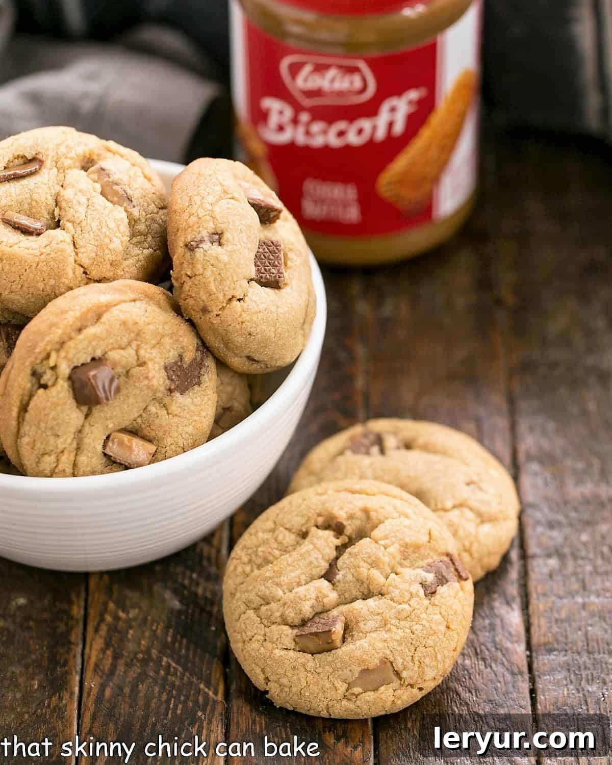 Two perfectly baked Toffee Biscoff Cookies resting next to a white bowl overflowing with more cookies, with a jar of Biscoff cookie butter in the background.