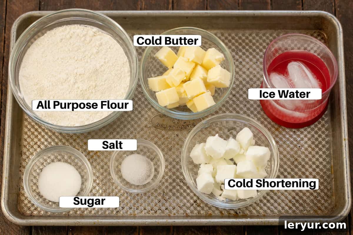 Pie Crust Ingredients with labels on a sheet pan. Essential ingredients for a perfect pie crust: flour, salt, sugar, cold cubed butter, cold cubed shortening, and a bowl of ice water.