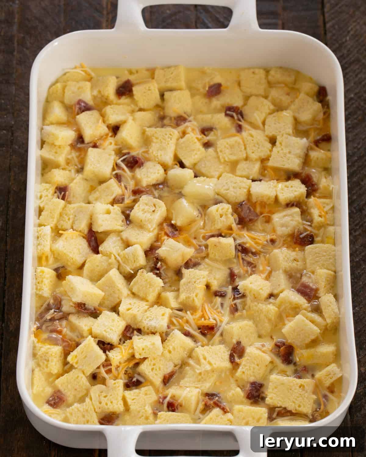 Cubed bread, bacon, and cheese in a baking dish, generously drizzled with melted butter and covered with egg custard.
