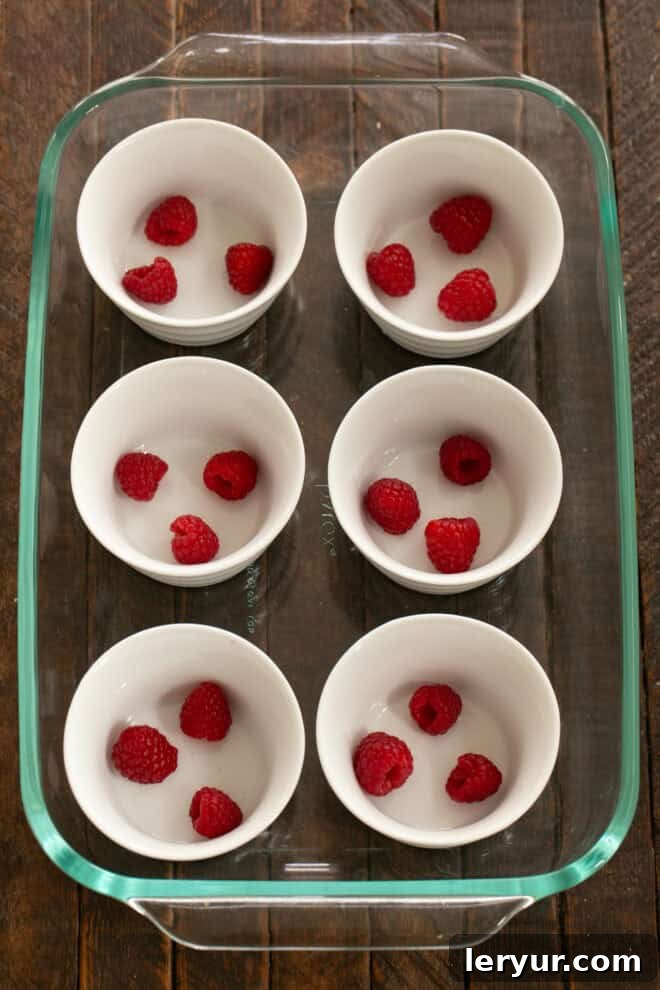 Six white ramekins arranged in a large rimmed baking pan, with fresh raspberries carefully placed at the bottom of each ramekin.