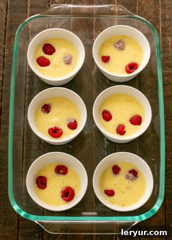 The lemon mixture being carefully poured into ramekins filled with raspberries, positioned in a baking pan, with warm water added to create a water bath.