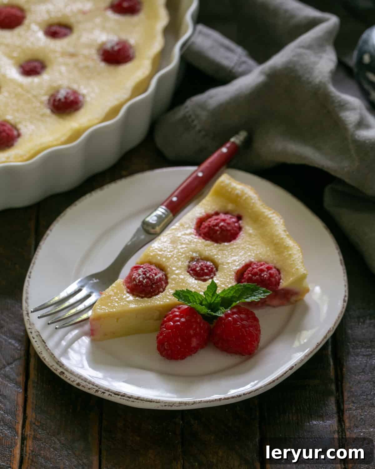 A perfectly portioned slice of raspberry clafoutis on a white dessert plate, accompanied by a fork with a red handle. The serving dish, still filled with more clafoutis, is visible in the soft-focused background.