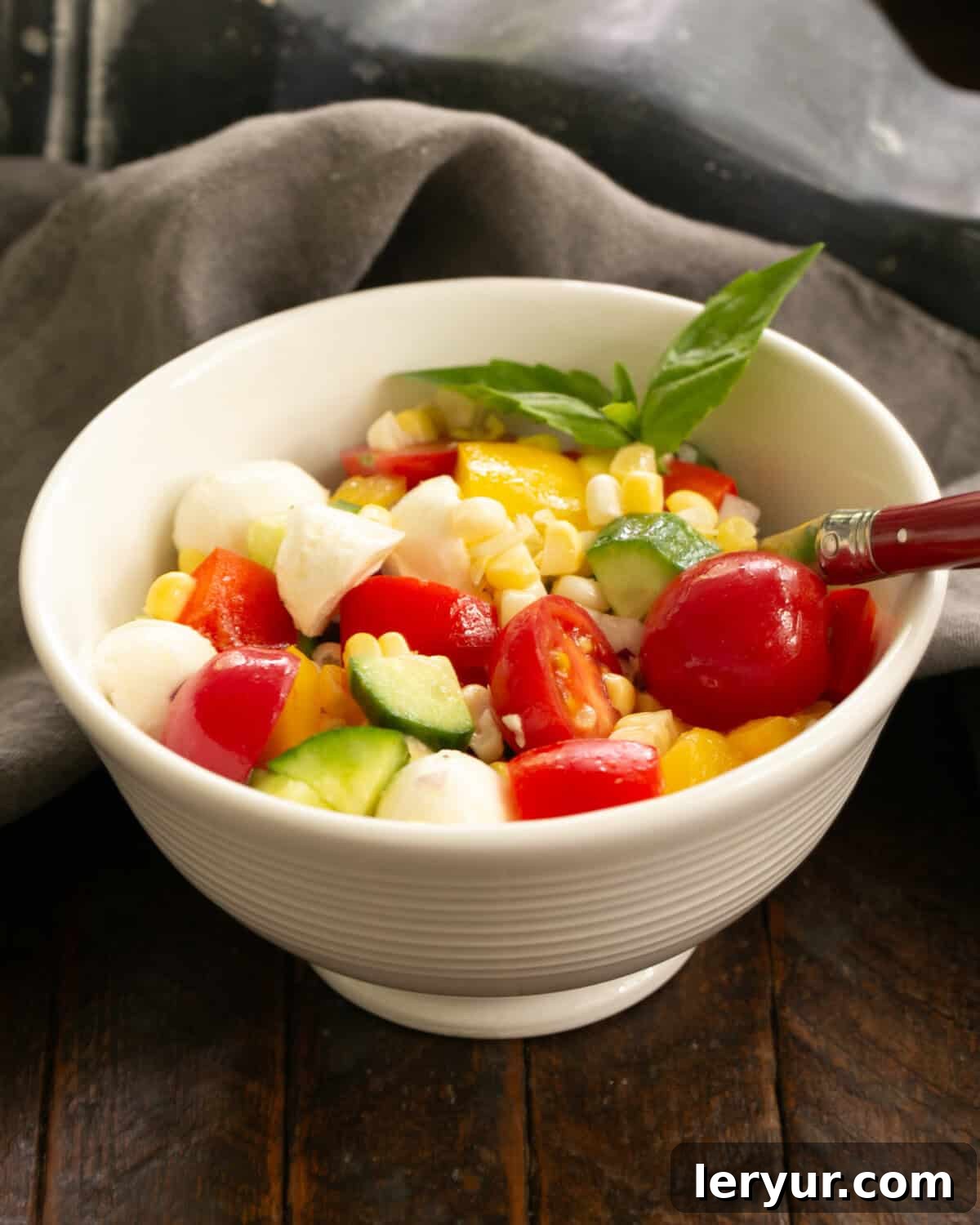 Kaleidoscope Summer Salad 2 A vibrant serving of Rainbow Summer Salad in a pristine white bowl, accompanied by a red-handled fork, showcasing colorful corn, tomatoes, bell peppers, and mozzarella.