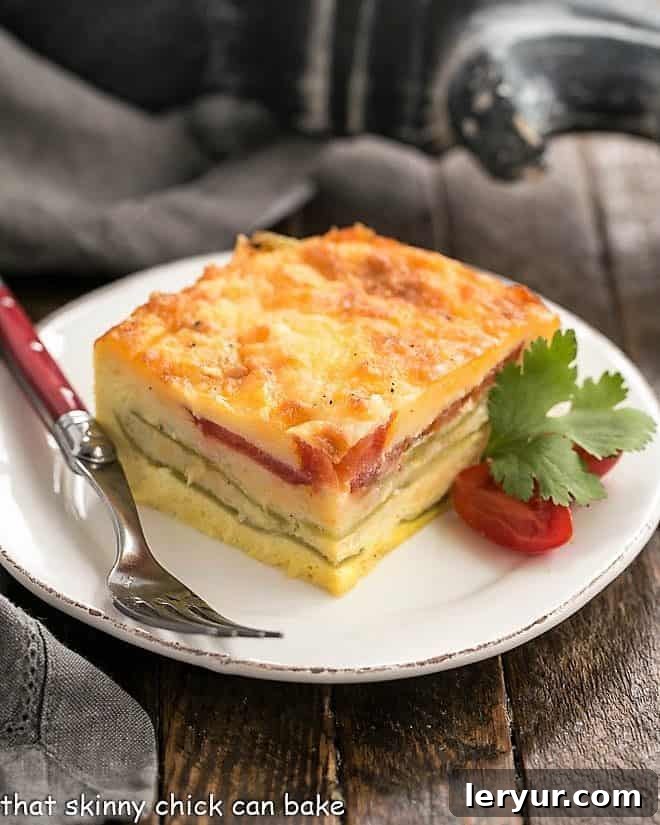 A perfectly portioned slice of Chile Relleno Casserole on a white plate, garnished with fresh cherry tomatoes and a sprig of cilantro, showcasing gooey layers of cheese, eggs, and green chiles.