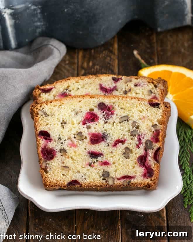 Classic Cranberry Pecan Bread is perfect for the holidays. The sweet-tart, crunchy-crispy components of this cranberry quick bread recipe make this a loaf to remember! Two vibrant slices of cranberry bread elegantly arranged on a white plate, garnished with a fresh orange slice for a pop of color.