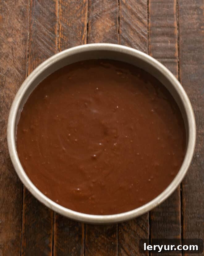 The smooth fudge cake batter being scraped into the prepared round cake pan.