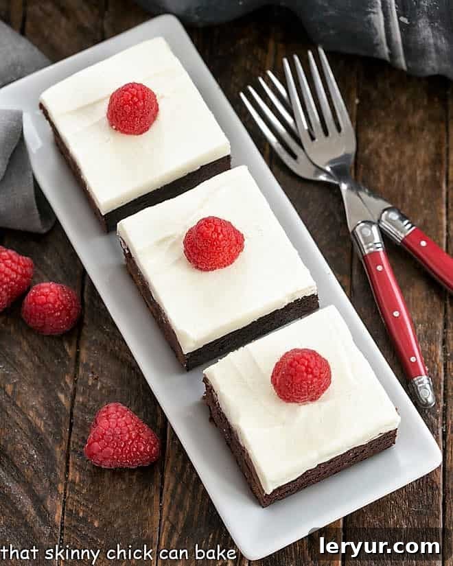 Overhead view of fudgy Raspberry Brownies with a swirl of creamy White Chocolate Frosting, artfully arranged on a pristine white serving tray, showcasing their rich color and inviting texture.