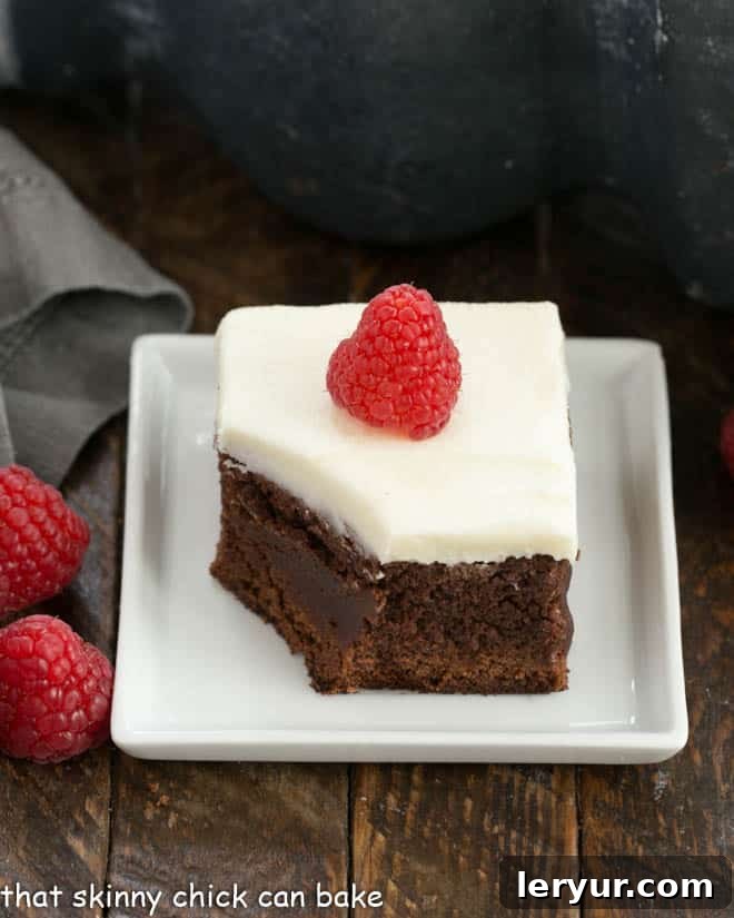 A perfectly portioned Raspberry Chambord Brownie, richly frosted with white chocolate cream cheese icing, sits on a square white plate, with a single bite removed to reveal its dense, fudgy interior and hidden raspberry flecks.