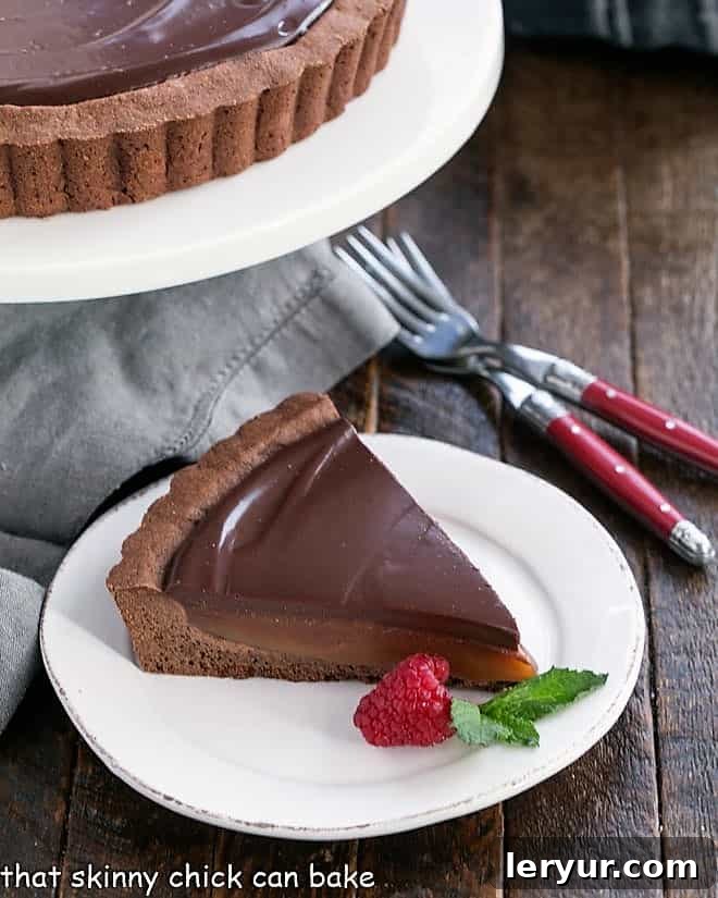 Chocolate Caramel Tart - A sublime pairing of chocolate and caramel makes for an outrageously delicious caramel dessert! A beautifully presented slice of Chocolate Caramel Tart on a white plate, accompanied by a cake stand and two forks.