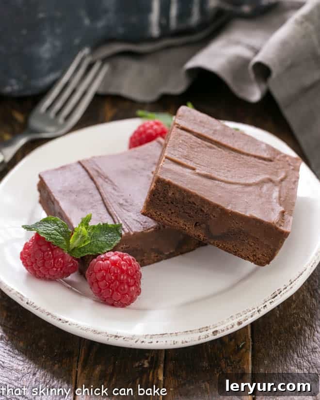 Chocolate Poke Brownies - The ganache seeps INTO the brownies!!! Two chocolate poke brownies artfully arranged on a white dessert plate, garnished with fresh raspberries and a sprig of mint, highlighting their rich, fudgy texture and glossy ganache topping.