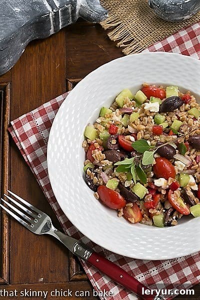 Delicious Greek Farro Salad with chewy farro, tomatoes, olives, cucumbers and more! Overhead view of Greek Farro Salad in a white bowl on a red and white checked napkin, showcasing its vibrant freshness.