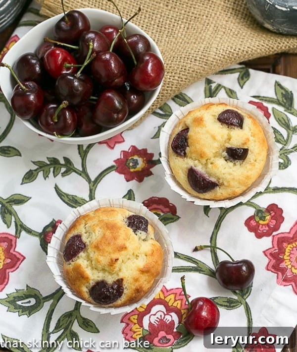 Two Double Cherry Muffins showcased on a charming floral napkin, with a rustic white bowl overflowing with dark, ripe cherries providing a beautiful backdrop, emphasizing the natural goodness.
