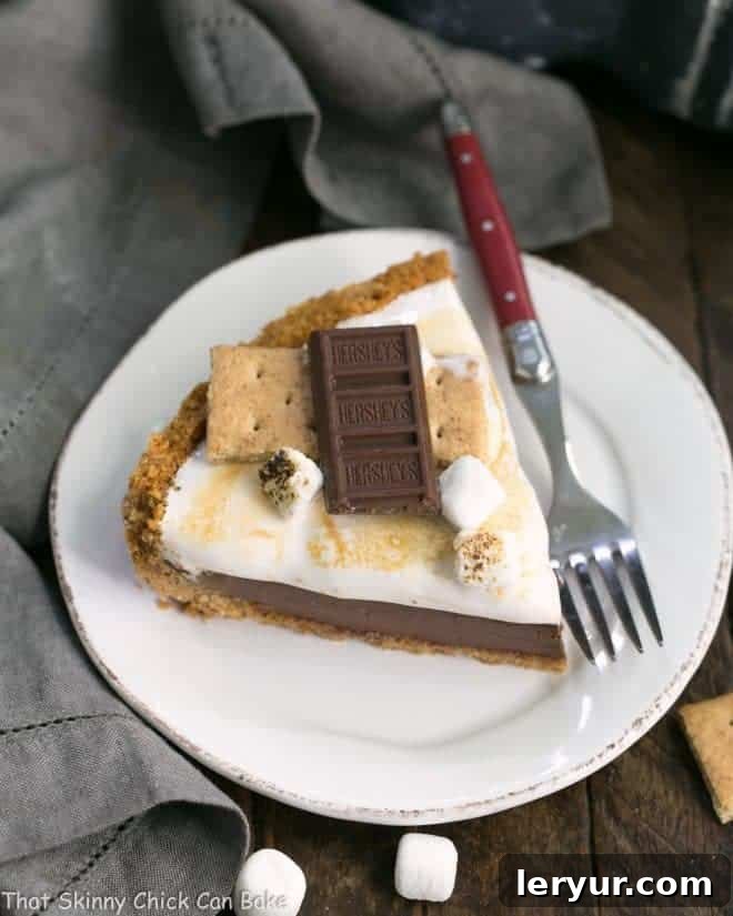 A perfectly sliced S'mores Pie on a white dessert plate, showcasing the distinct layers of graham cracker crust, chocolate ganache, and fluffy toasted marshmallow topping.