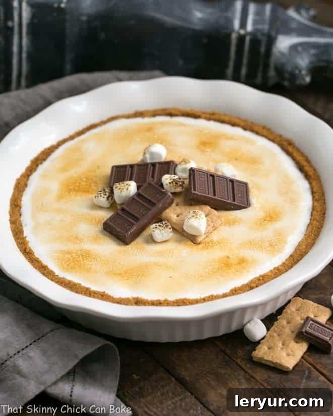A whole S'mores Pie, garnished with chocolate pieces and miniature marshmallows, presented in a white pie plate. The toasted marshmallow topping is golden and inviting.
