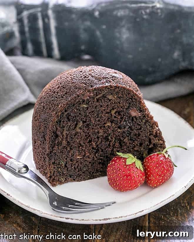 A perfectly sliced piece of Chocolate Zucchini Bundt Cake on a white plate, garnished with a red-handled fork and two fresh strawberries.