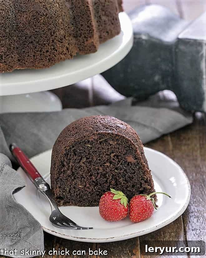A moist slice of zucchini cake on a white plate, with the entire Bundt cake in the background on a pristine white cake stand.