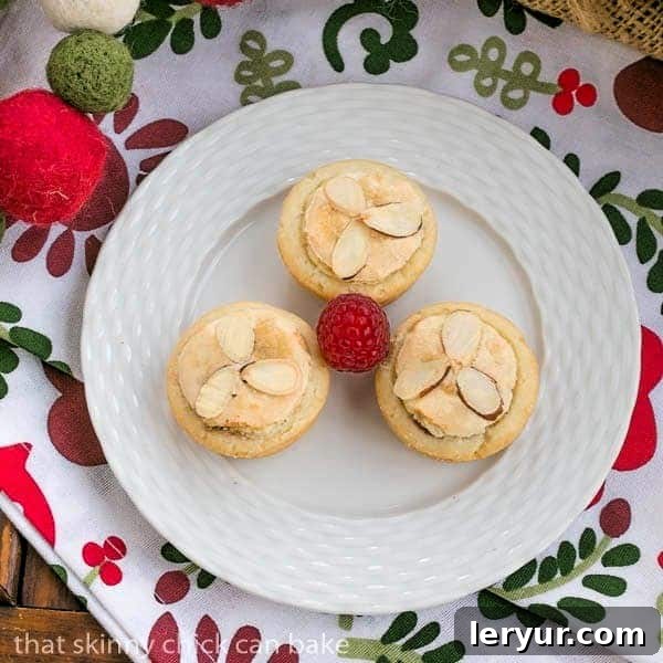Three Almond Raspberry Tartlets on a white plate, garnished with fresh raspberries and a dusting of powdered sugar.