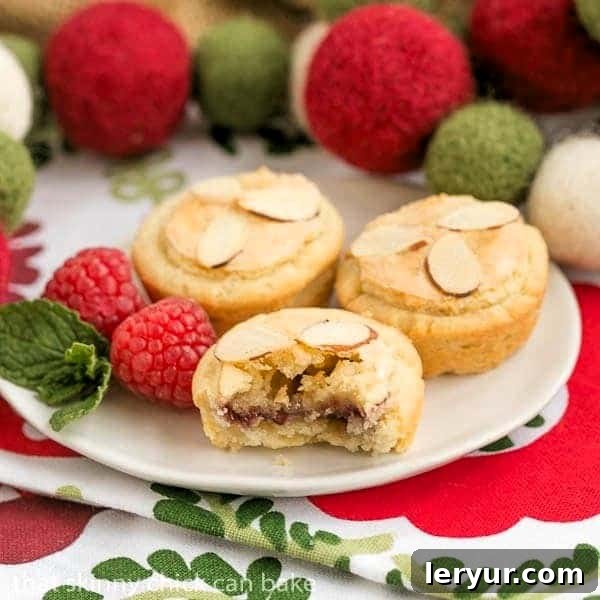 Close-up of Almond Raspberry Tartlets on a white plate, one tartlet cut in half to show the distinct layers of raspberry jam and almond paste filling.