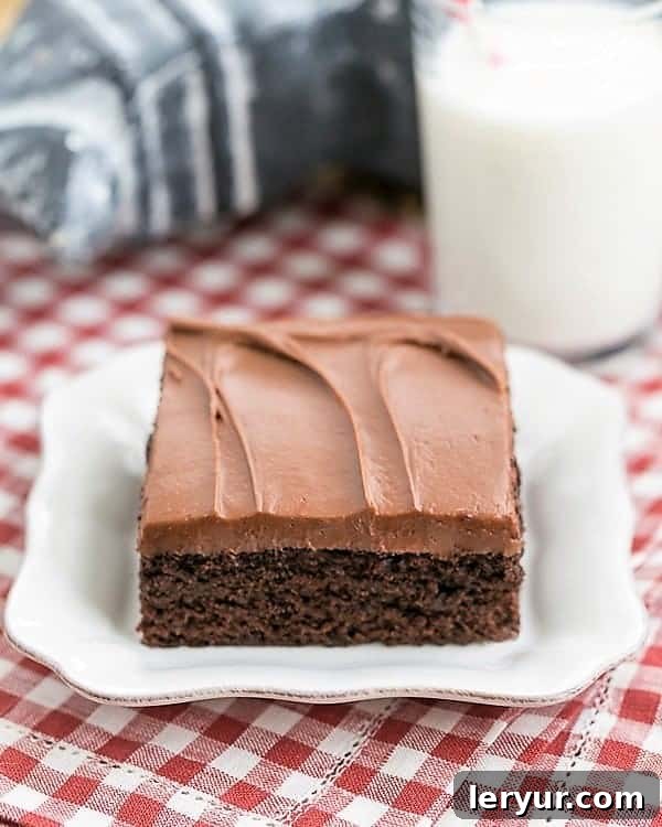 Frosted Fudge Cake Recipe - Easy and outrageously delicious! Cocoa Fudge Cake on a square white plate with a glass of milk, highlighting its rich, dark chocolate color and creamy frosting.