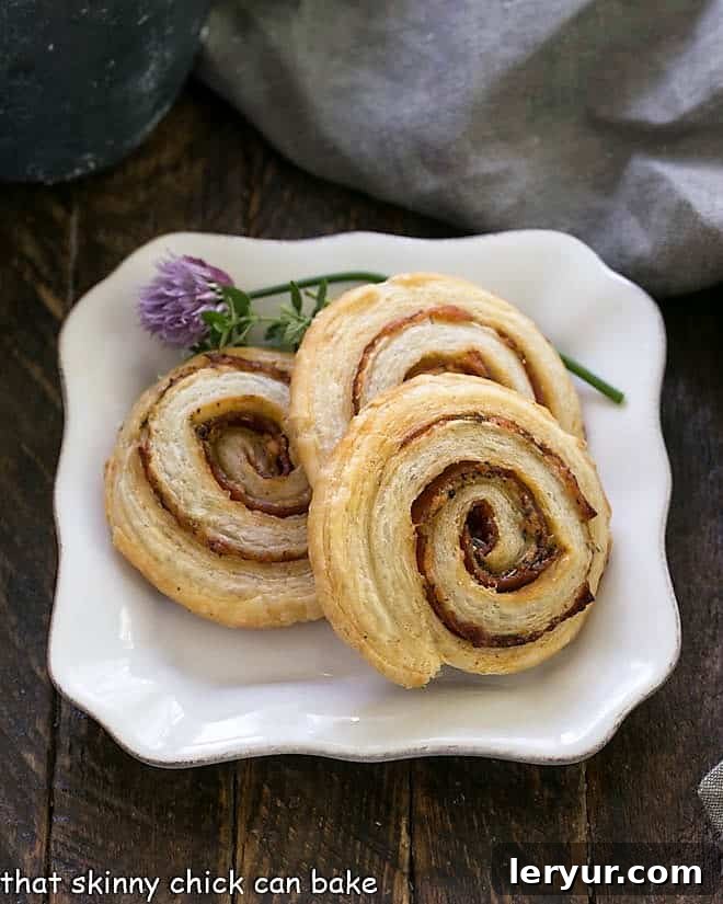 Close-up of golden Honey Mustard Pepperoni Pinwheels on a square white plate, garnished with vibrant green flowering chives, highlighting their flaky texture and savory filling.