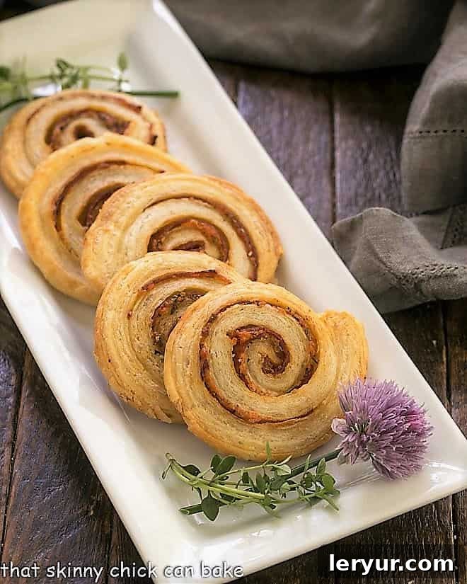 Three golden-brown Honey Mustard Pepperoni Pinwheels displayed on a white ceramic tray, with a blossoming chive adding a fresh accent. The flaky layers of the pastry are clearly visible.