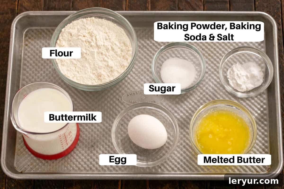 Fluffy Buttermilk Pancakes 2 A flat lay photograph of various pancake ingredients neatly arranged on a silver sheet pan, including a bowl of flour, sugar, salt, baking powder, baking soda, a carton of buttermilk, an egg, and a stick of butter, all clearly labeled.