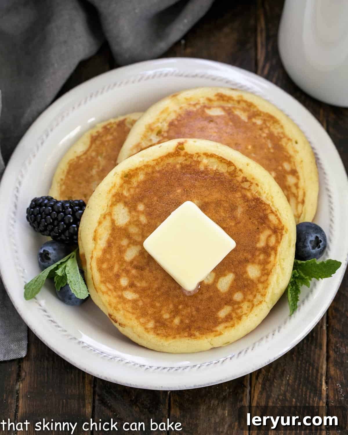 Fluffy Buttermilk Pancakes -This easy breakfast recipe trumps a box mix every time. A breeze to make with ingredients you'll already find in your kitchen. Overhead view of three perfectly cooked, golden-brown buttermilk pancakes, stacked and generously topped with a dollop of melting butter, garnished with fresh mixed berries and a sprig of mint for a vibrant touch.